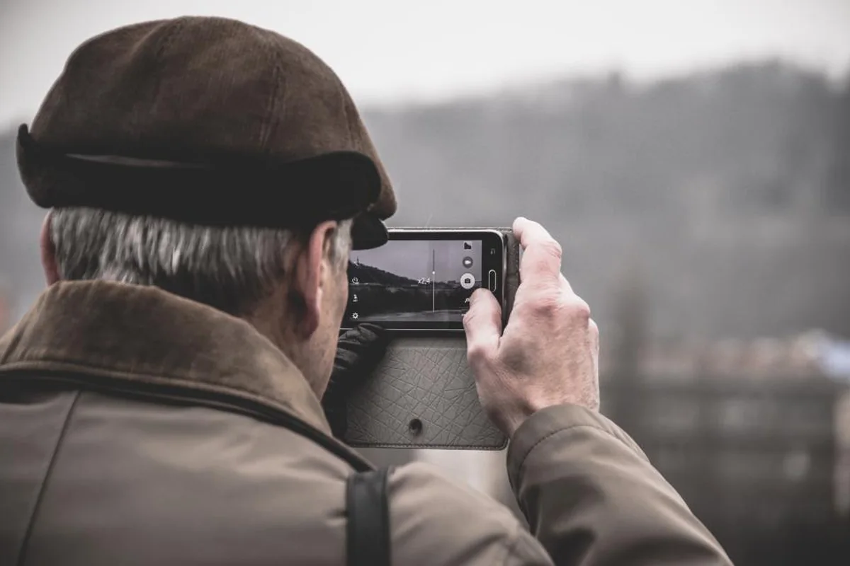 Hombre mayor con gorra y abrigo marrón, de espaldas, sujetando un móvil con ambas manos para fotografiar un paisaje de montaña con niebla