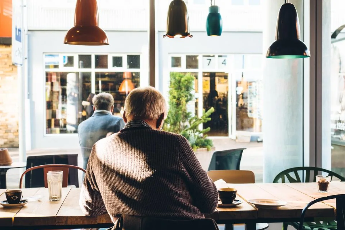 Un hombre mayor de espaldas, sentado solo en la mesa de una cafetería junto a un gran ventanal