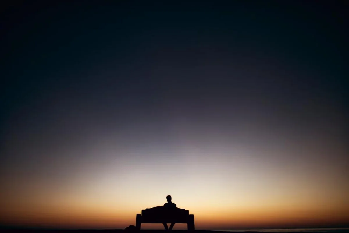 Silueta de una persona sentada sola en un banco frente al horizonte al atardecer, con el cielo pasando del naranja al azul oscuro