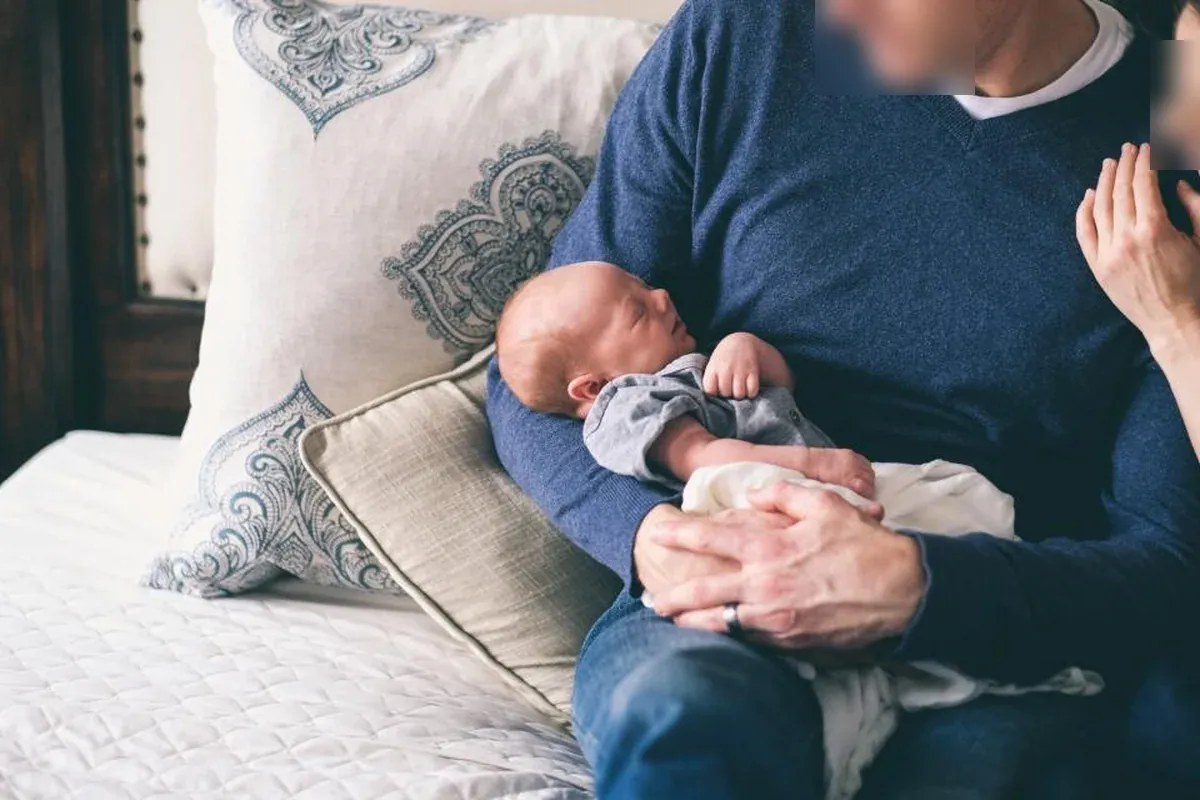 Hombre con jersey azul sosteniendo a un recién nacido dormido sobre una cama con cojines, mientras otra persona apoya la mano en su hombro