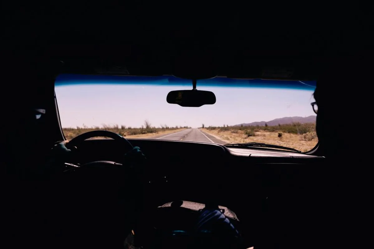 Interior de un coche en penumbra con el volante y la silueta del conductor, mirando a través del parabrisas una carretera recta entre campos