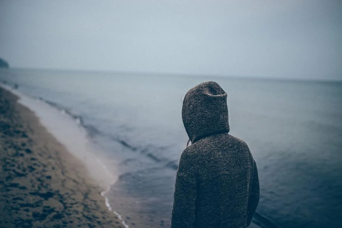 Joven con capucha de punto, de espaldas, mirando el mar en una playa de día nublado