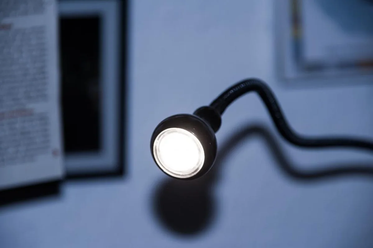 Pequeña lámpara de escritorio con brazo flexible encendida en una habitación en penumbra, con un libro abierto desenfocado al fondo