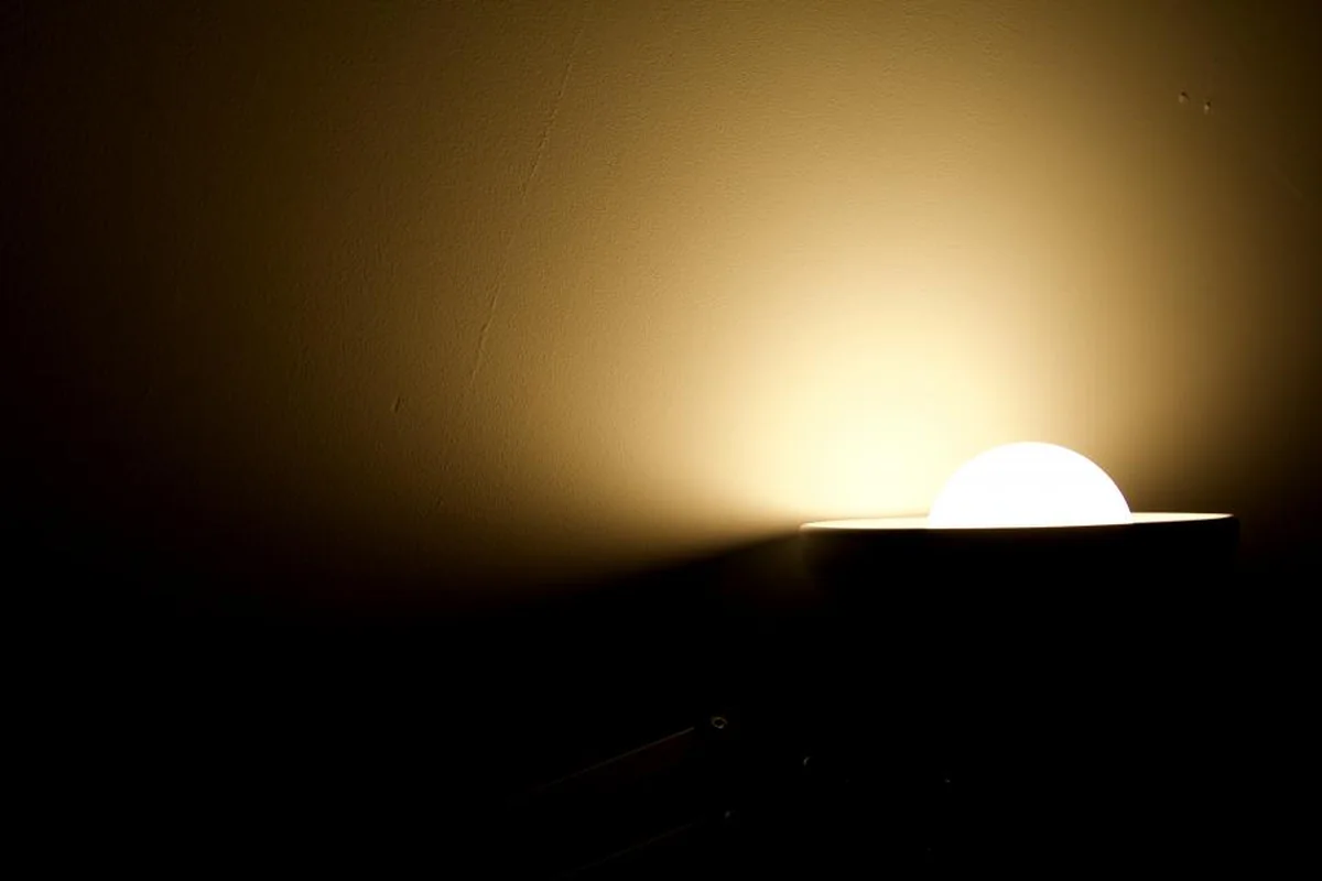Lámpara de luz cálida encendida sobre un mueble en una habitación casi a oscuras, con la pared iluminada en tono ámbar