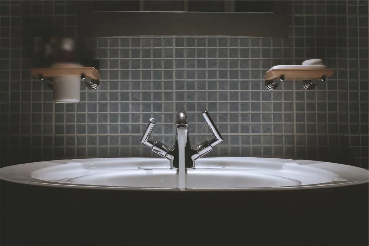 Lavabo blanco iluminado en un baño oscuro con azulejos pequeños y dos jaboneras de madera