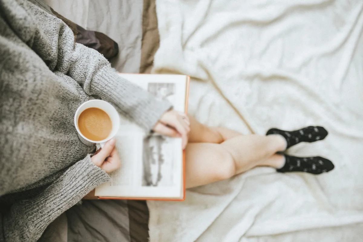 Persona con jersey gris leyendo un libro sobre la cama mientras sujeta una taza de café con leche