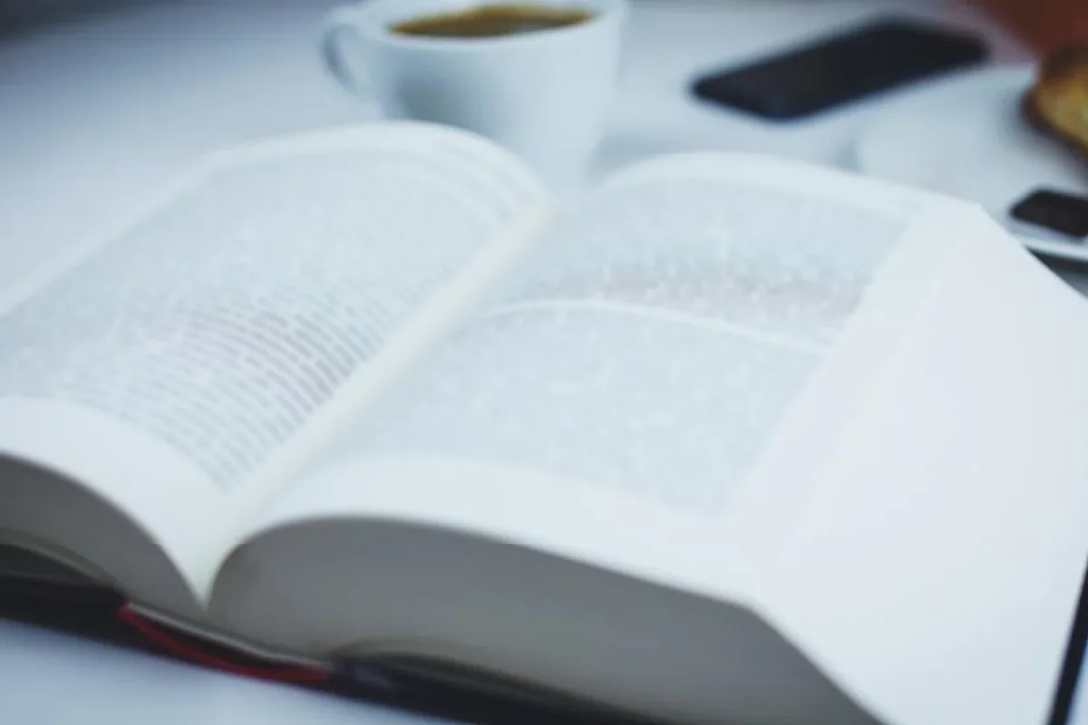 Libro grueso abierto sobre una mesa blanca, con una taza de café y un teléfono móvil desenfocados al fondo