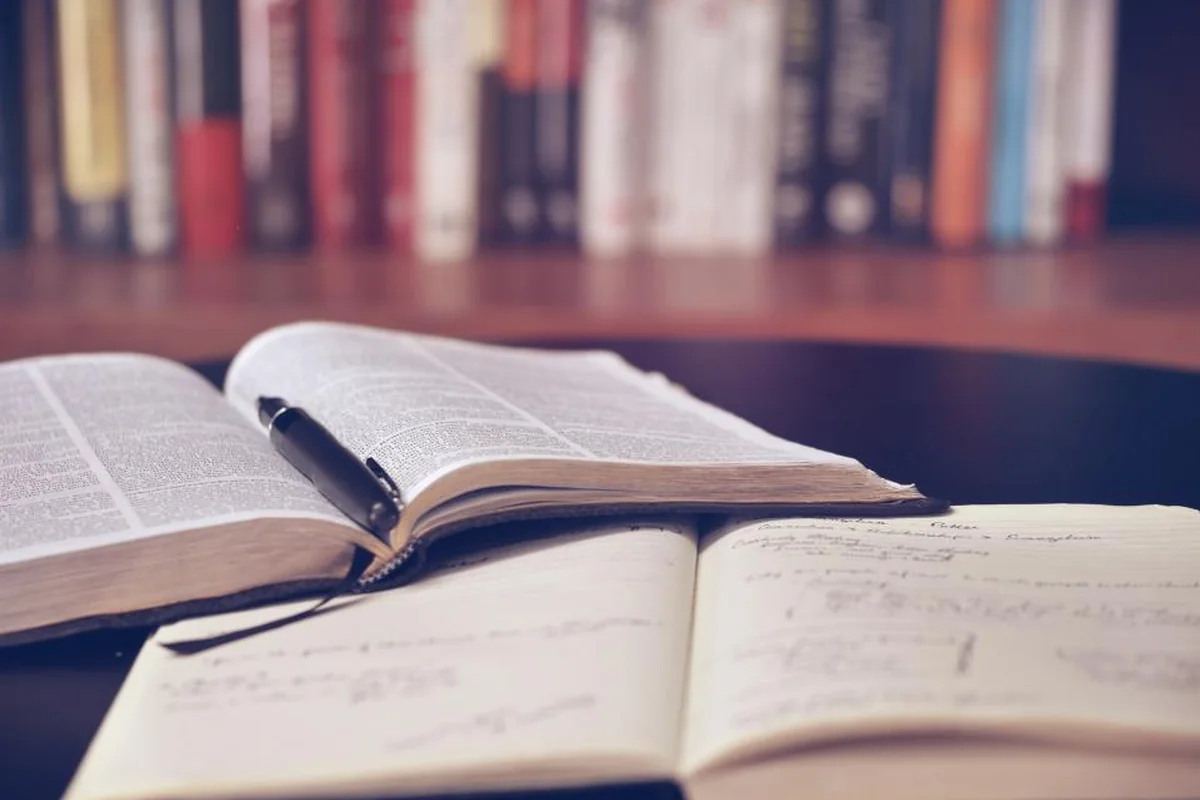 Dos libros abiertos con un bolígrafo negro y apuntes a mano sobre un escritorio, con una estantería llena de libros al fondo