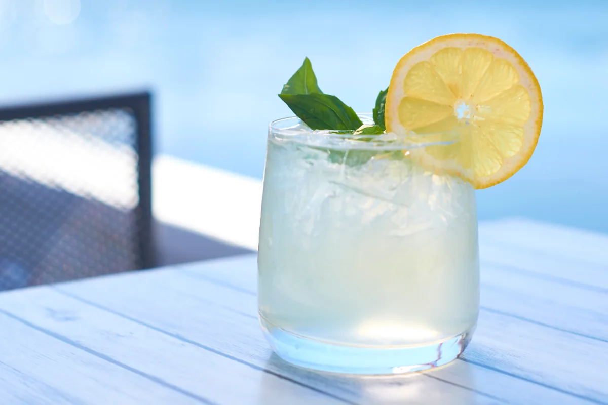 Vaso de limonada con hielo, una rodaja de limón y hojas de menta sobre una mesa de madera blanca en una terraza soleada