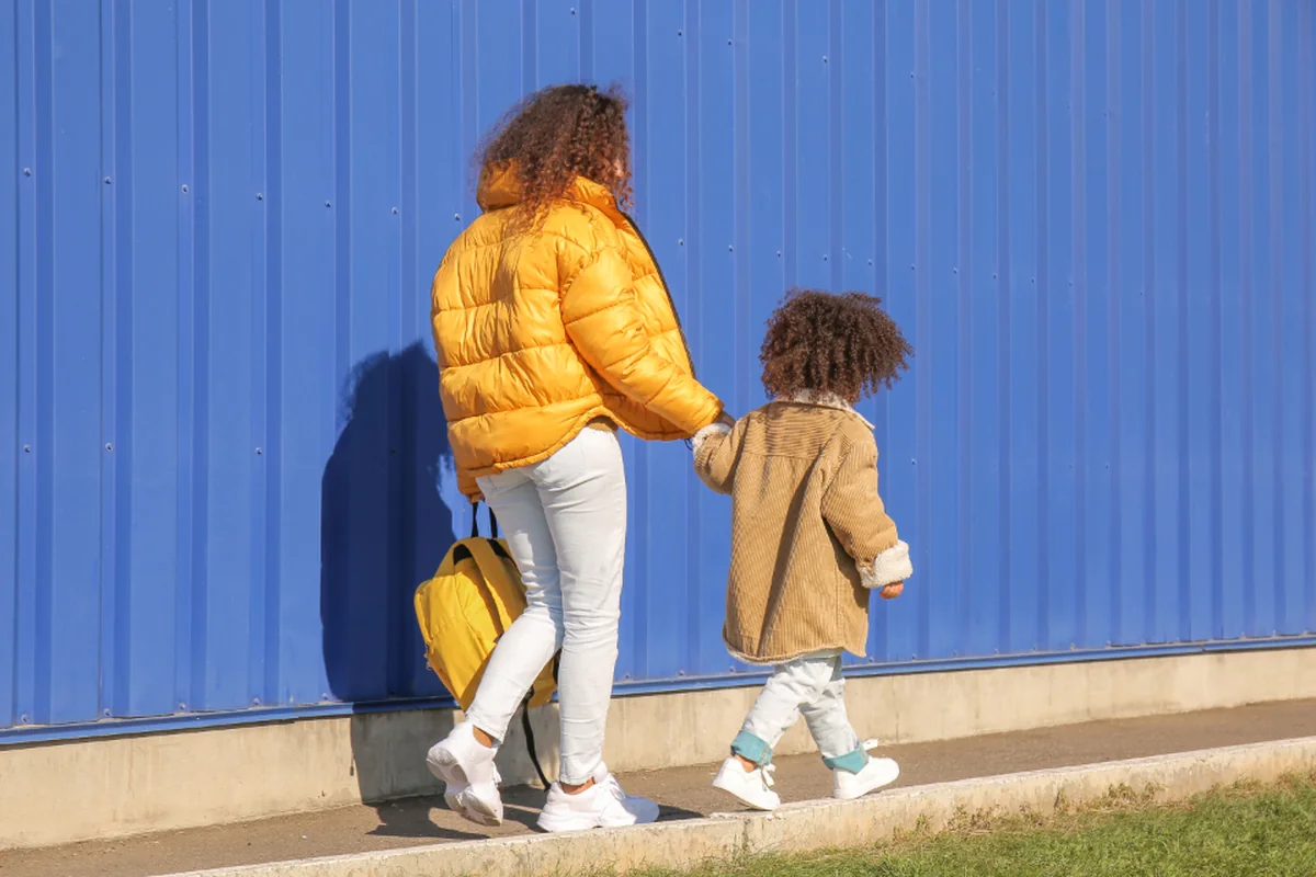 Una mujer con chaqueta amarilla y un niño pequeño caminan de espaldas, de la mano, junto a un muro azul