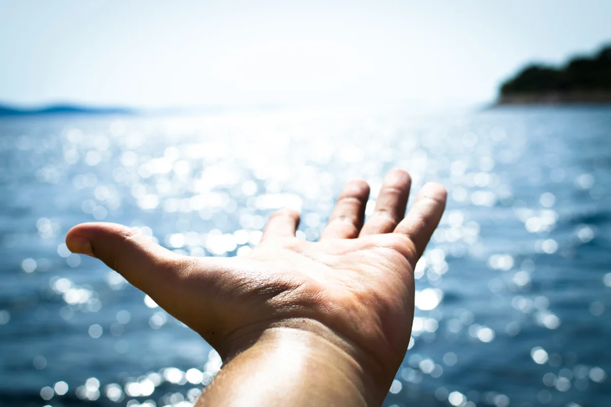 Mano abierta extendida hacia el mar con reflejos de sol sobre el agua y una costa al fondo