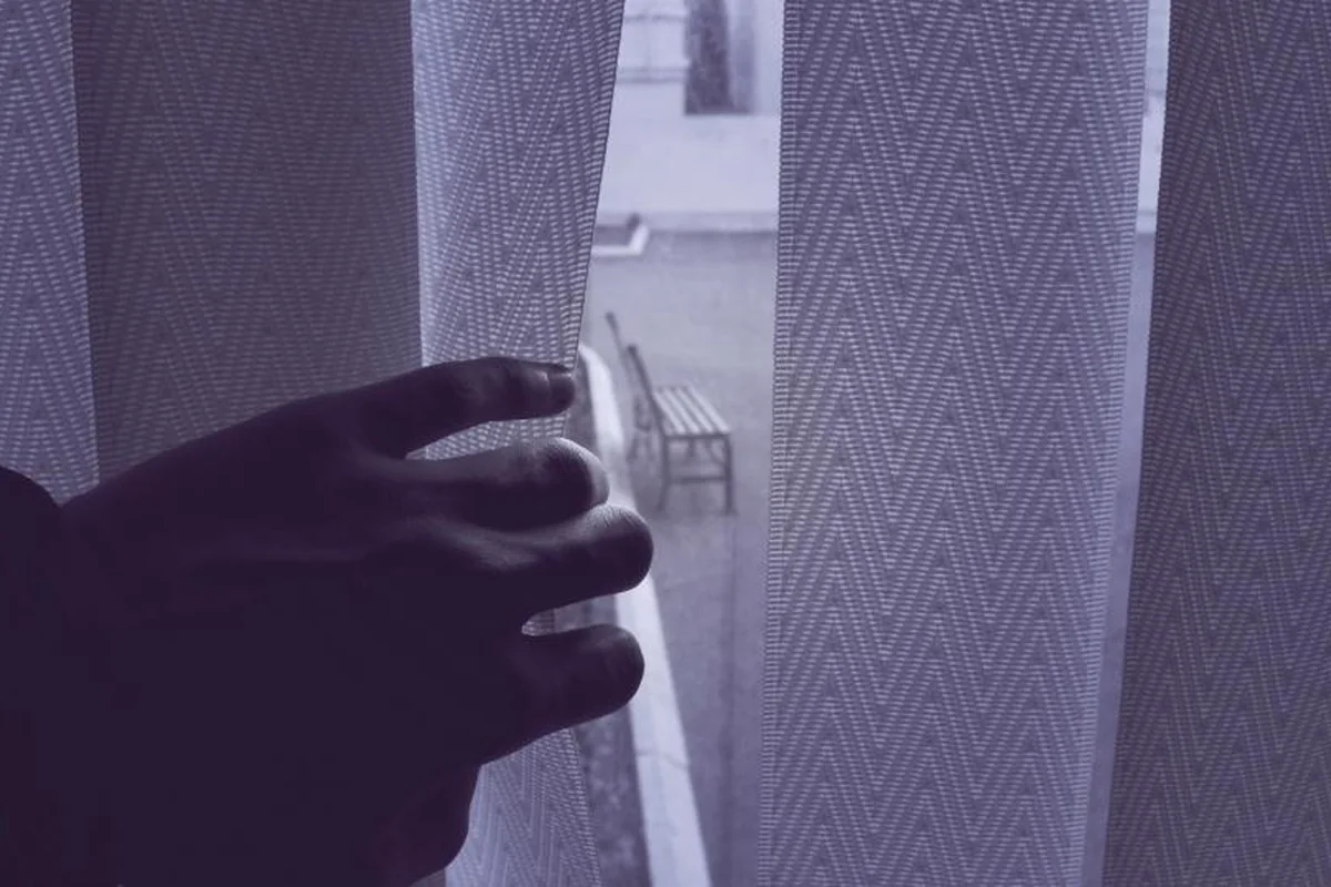 Mano que aparta una cortina de tela gris para mirar a través de la ventana, donde se intuye un banco en la calle