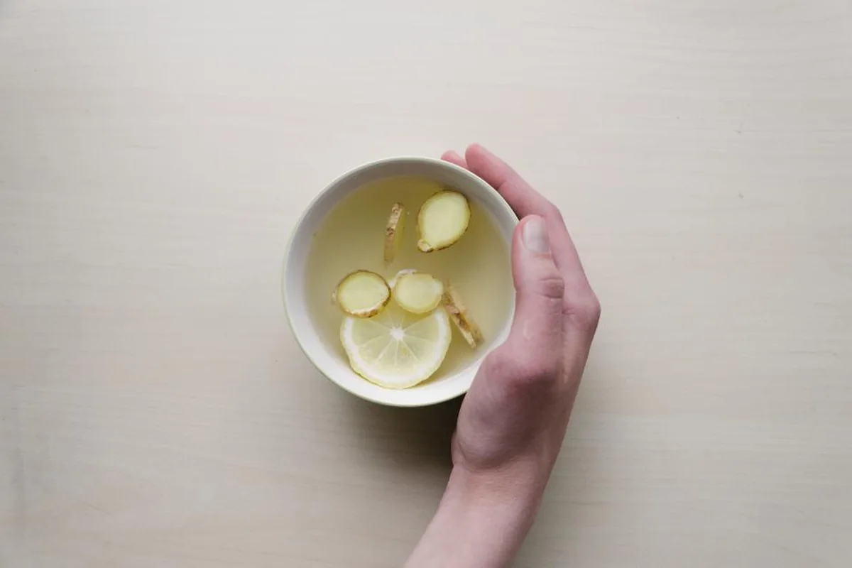 Mano sujetando un cuenco blanco con una infusión de jengibre y una rodaja de limón sobre una mesa clara