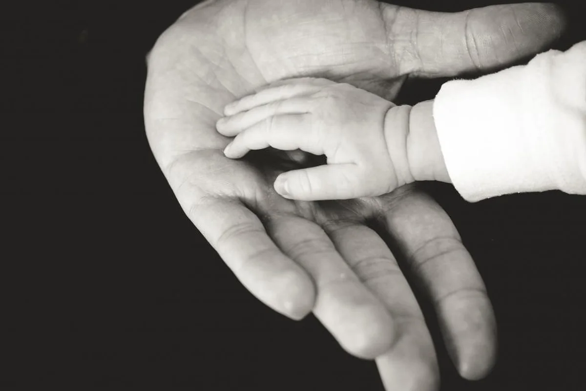 Fotografía en blanco y negro de la mano abierta de un adulto sobre la que descansa la pequeña mano de un bebé con la manga de un pijama