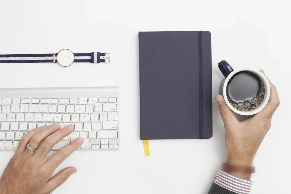 Una mano sobre un teclado blanco y otra sujetando una taza de café, junto a una libreta gris y un reloj de pulsera