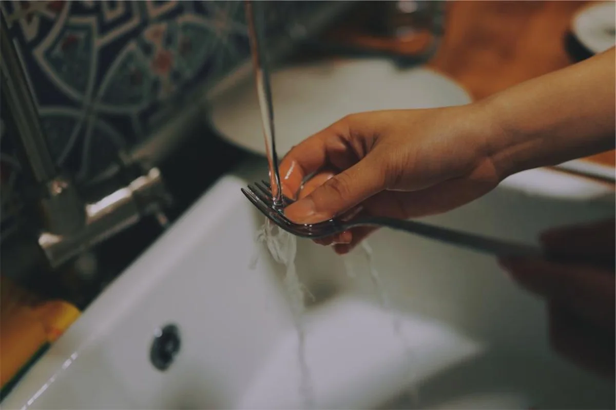 Mano enjuagando un tenedor bajo el chorro del grifo sobre un fregadero blanco, con azulejos estampados al fondo