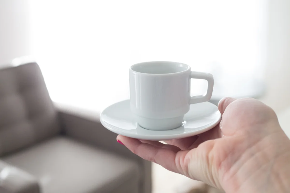 Mano que sostiene una taza blanca de café sobre su plato en un salón luminoso con un sofá gris al fondo