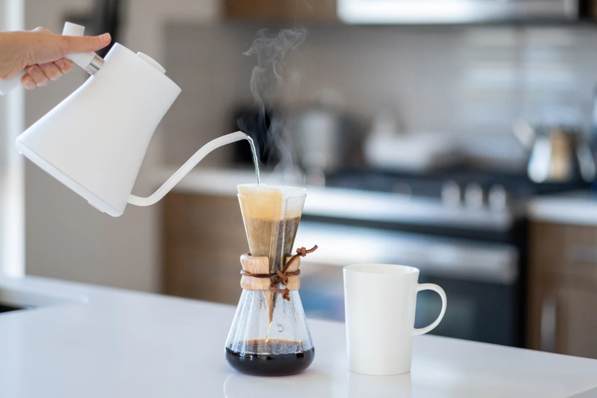 Mano que vierte agua caliente con un hervidor blanco sobre una cafetera de filtro, junto a una taza en la encimera de una cocina