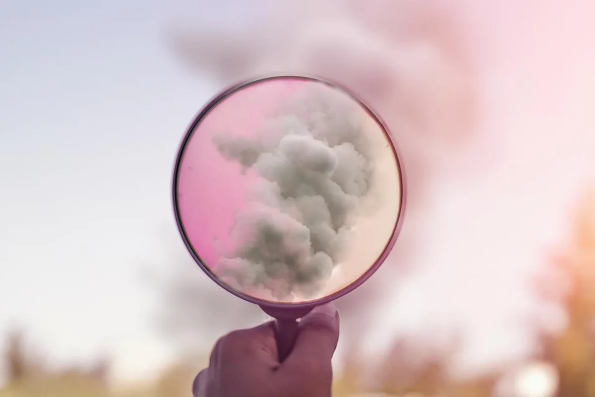 Mano sosteniendo un espejo redondo que refleja una nube de humo rosa sobre un fondo desenfocado