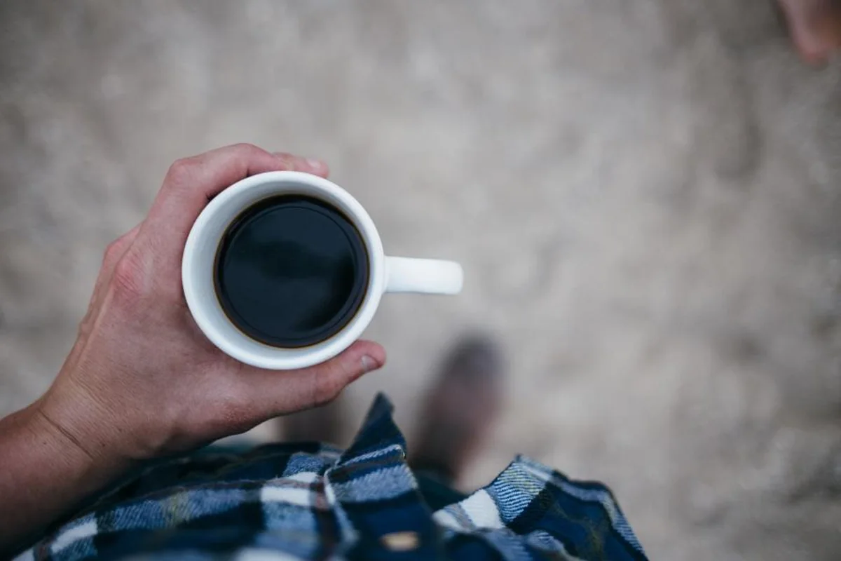 Mano sosteniendo una taza blanca de café solo, vista desde arriba, con una camisa de cuadros y el suelo desenfocado al fondo