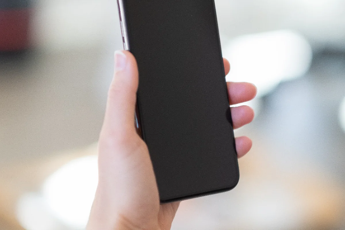 Mano sosteniendo un teléfono móvil con la pantalla apagada sobre un fondo luminoso desenfocado