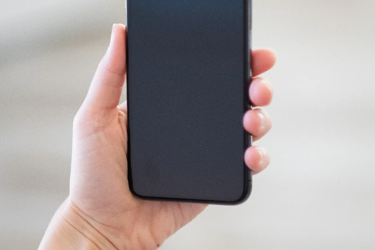 Mano de una persona sujetando un teléfono móvil con la pantalla apagada sobre un fondo claro desenfocado