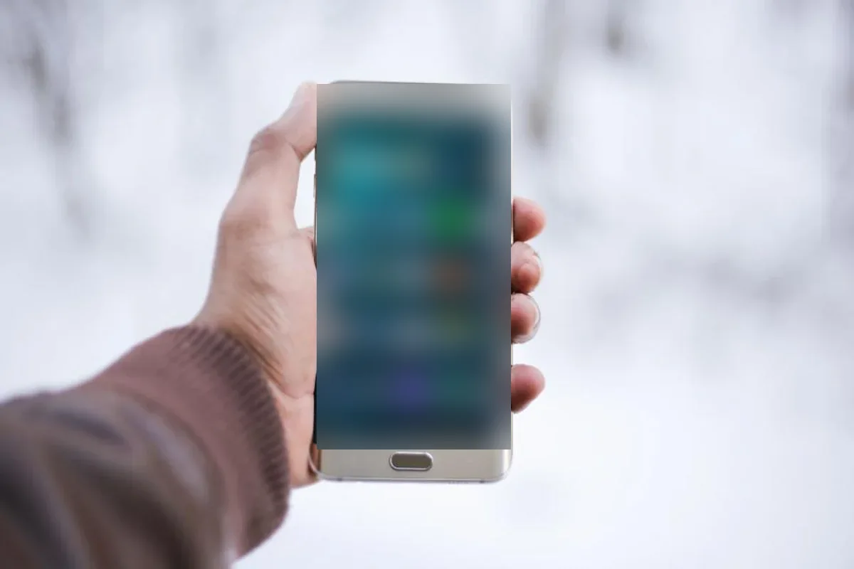 Mano que sostiene un teléfono móvil con la pantalla encendida y desenfocada, sobre un fondo blanco y nevado