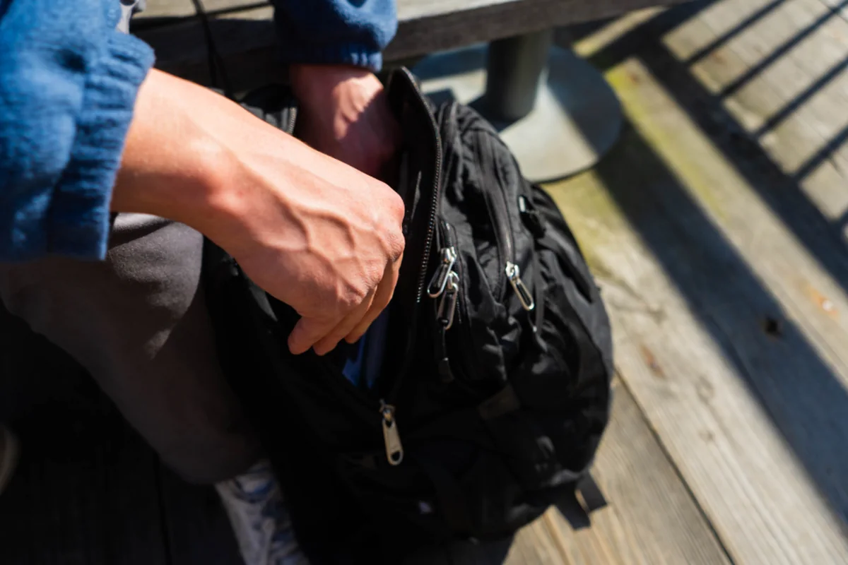 Unas manos abren la cremallera de una mochila negra apoyada sobre un banco de madera al sol