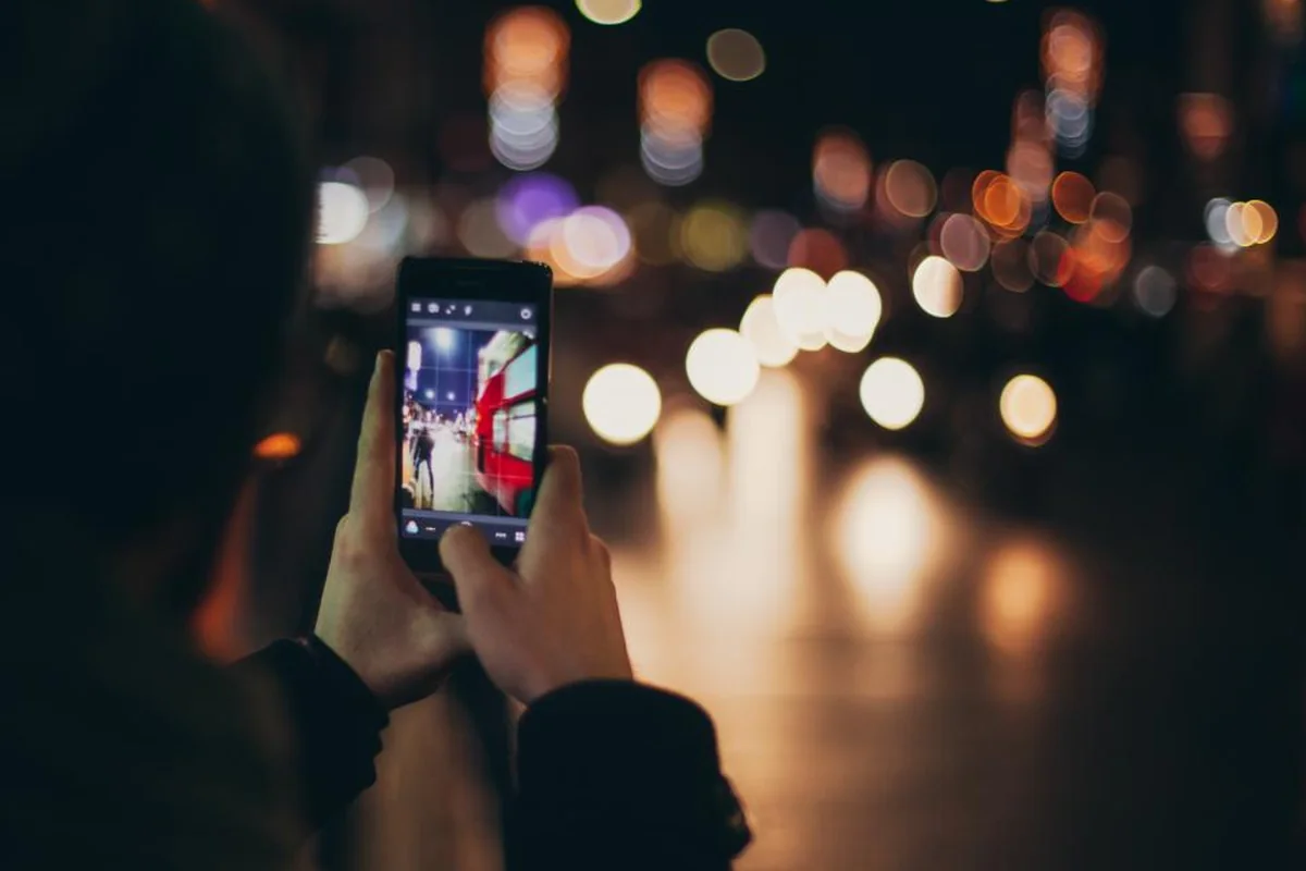 Manos sujetando un teléfono móvil de noche, con las luces de la ciudad desenfocadas al fondo