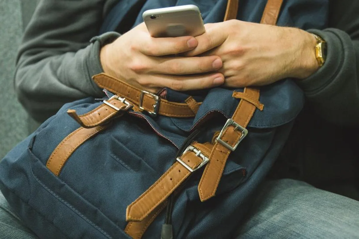 Manos de un joven sujetando un móvil sobre una mochila azul con correas de cuero