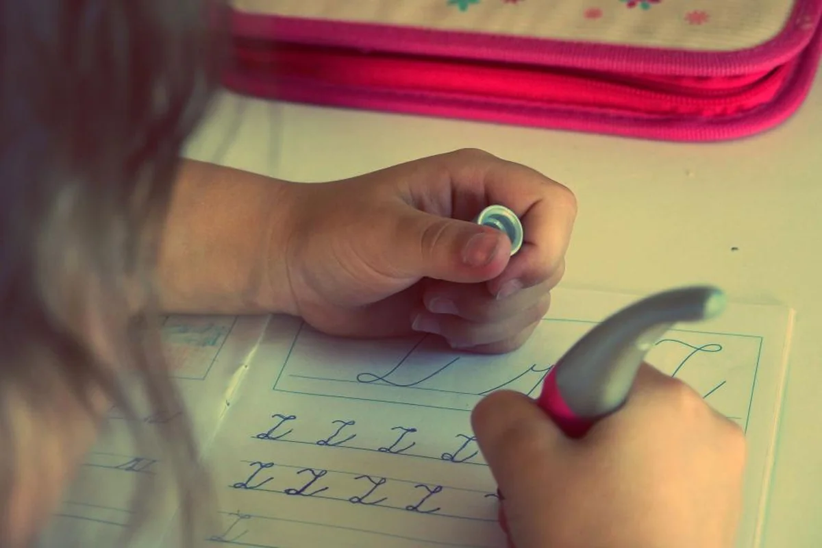 Manos de una niña escribiendo letras de caligrafía en un cuaderno junto a un estuche rosa