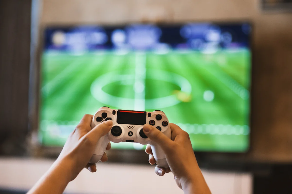 Manos sujetando un mando de consola blanco frente a un televisor con un videojuego de fútbol