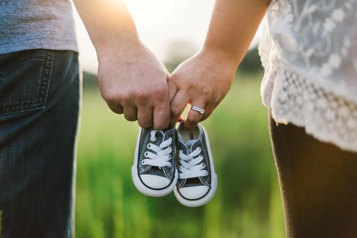 Dos personas cogidas de la mano sosteniendo un par de zapatillas de bebé grises, con un prado verde al fondo