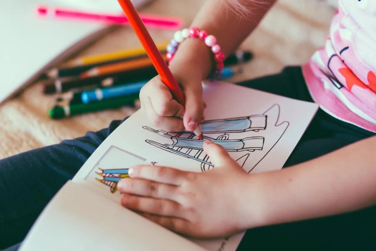 Las manos de una niña con una pulsera de cuentas colorean un dibujo con un lápiz rojo, rodeadas de lápices de colores
