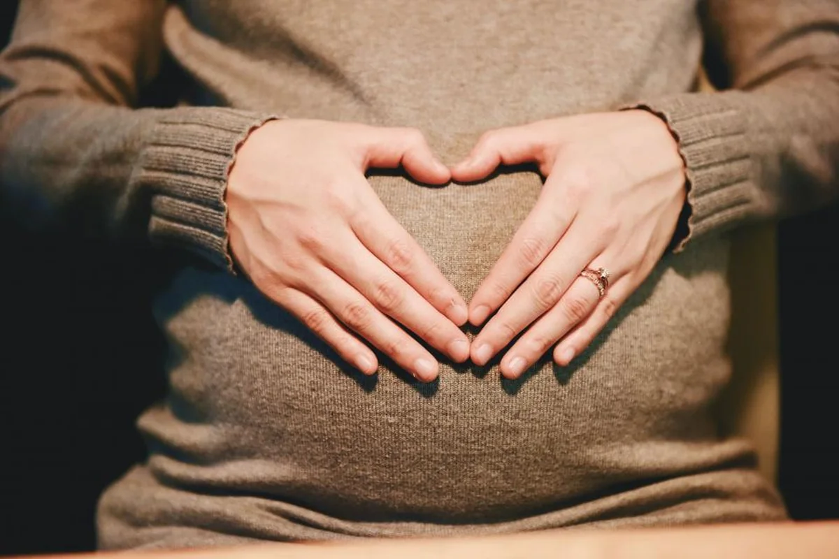 Manos formando un corazón sobre el vientre de una mujer embarazada con jersey marrón