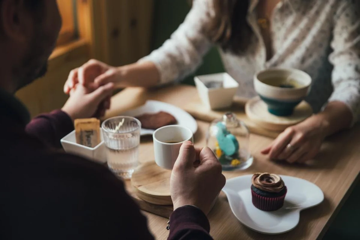 Dos personas se dan la mano sobre una mesa de cafetería con tazas, vasos de agua y un pastel