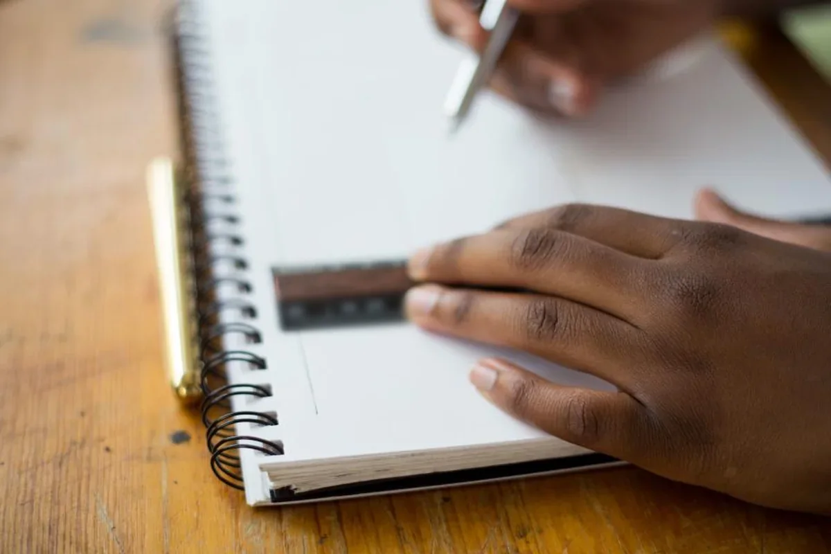 Manos apoyadas sobre un cuaderno de espiral con una regla mientras otra mano escribe, sobre una mesa de madera