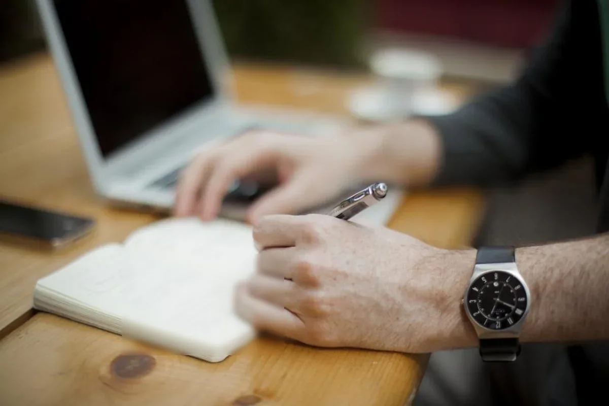 Manos de un hombre con reloj de pulsera escribiendo en una libreta abierta sobre una mesa de madera, junto a un portátil
