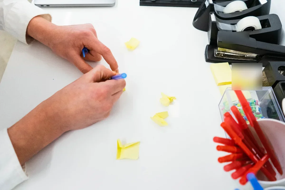 Manos escribiendo sobre pequeñas notas adhesivas amarillas en un escritorio blanco, junto a una grapadora, un portacelos y un vaso con rotuladores rojos