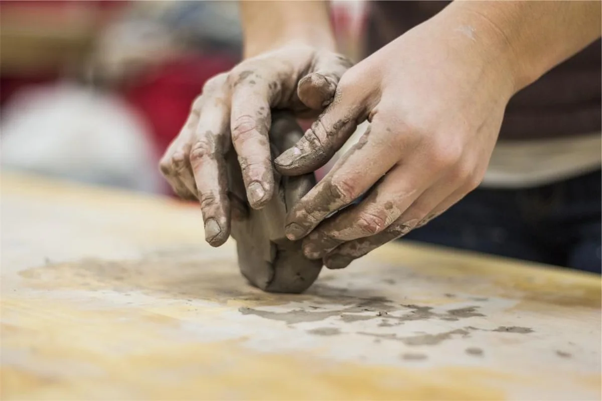 Dos manos manchadas de barro dando forma a un trozo de arcilla sobre una mesa de madera