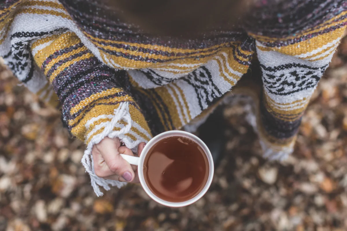 Vista cenital de una mano que sostiene una taza de té, con el brazo envuelto en una manta de rayas amarillas y grises sobre un suelo de hojas secas