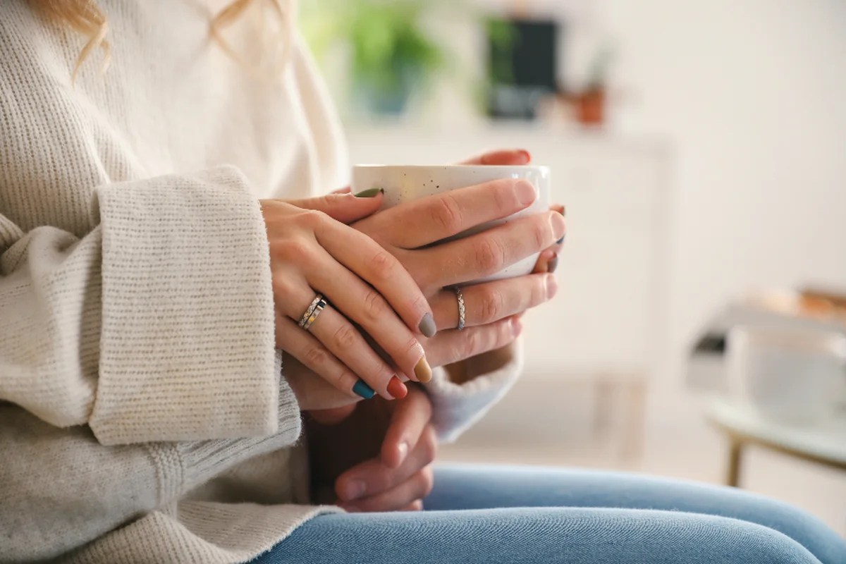 Manos de una persona con jersey de punto claro sujetando una taza blanca, sentada en un sofá