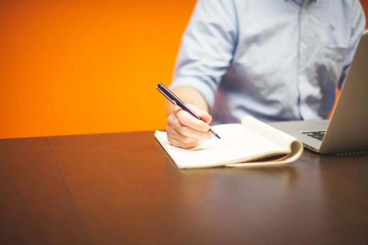 Persona con camisa azul toma notas a mano en una libreta junto a un portátil, sobre una mesa oscura y fondo naranja