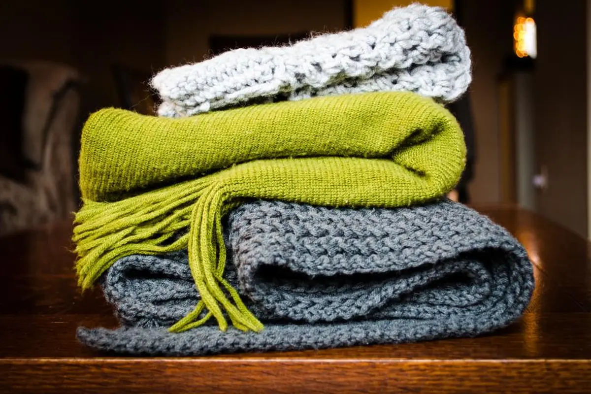 Tres mantas de punto dobladas y apiladas, una blanca, una verde con flecos y una gris, sobre una mesa de madera en una habitación con luz cálida