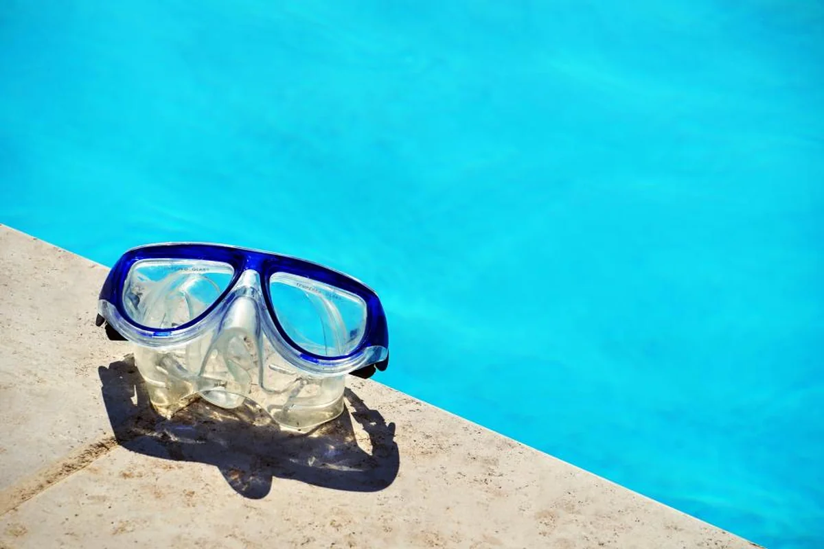 Máscara de buceo con montura azul apoyada en el borde de piedra de una piscina de agua turquesa