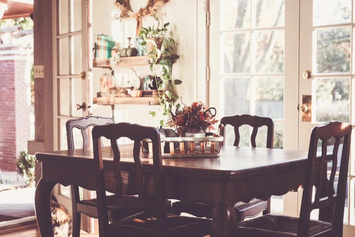 Mesa de comedor de madera con sillas vacías junto a unas puertas acristaladas y estanterías con plantas