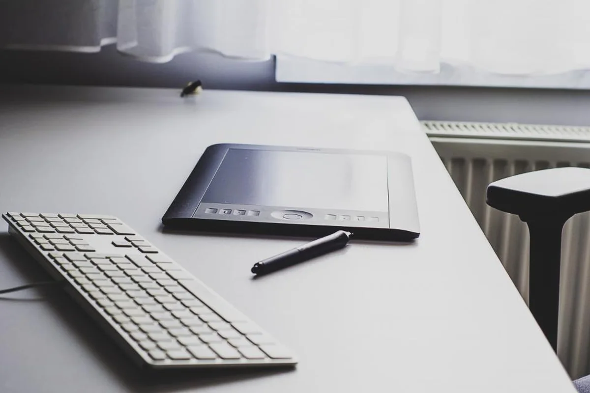 Mesa de trabajo blanca y ordenada con un teclado, una tableta gráfica y un lápiz junto a una ventana con cortina