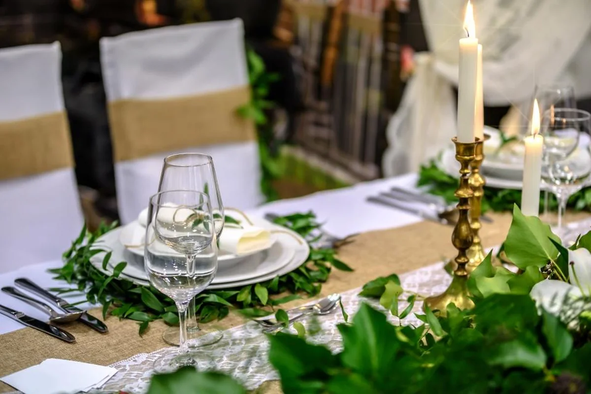 Mesa larga preparada para una celebración, con mantel de arpillera, copas, platos blancos, guirnalda de hojas verdes y velas encendidas en candelabros dorados