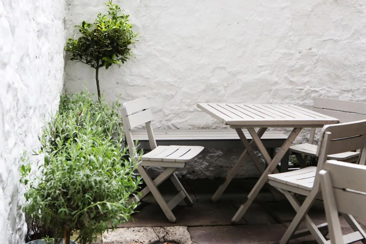 Una mesa de madera clara con sillas plegables vacías en un patio de paredes blancas con plantas
