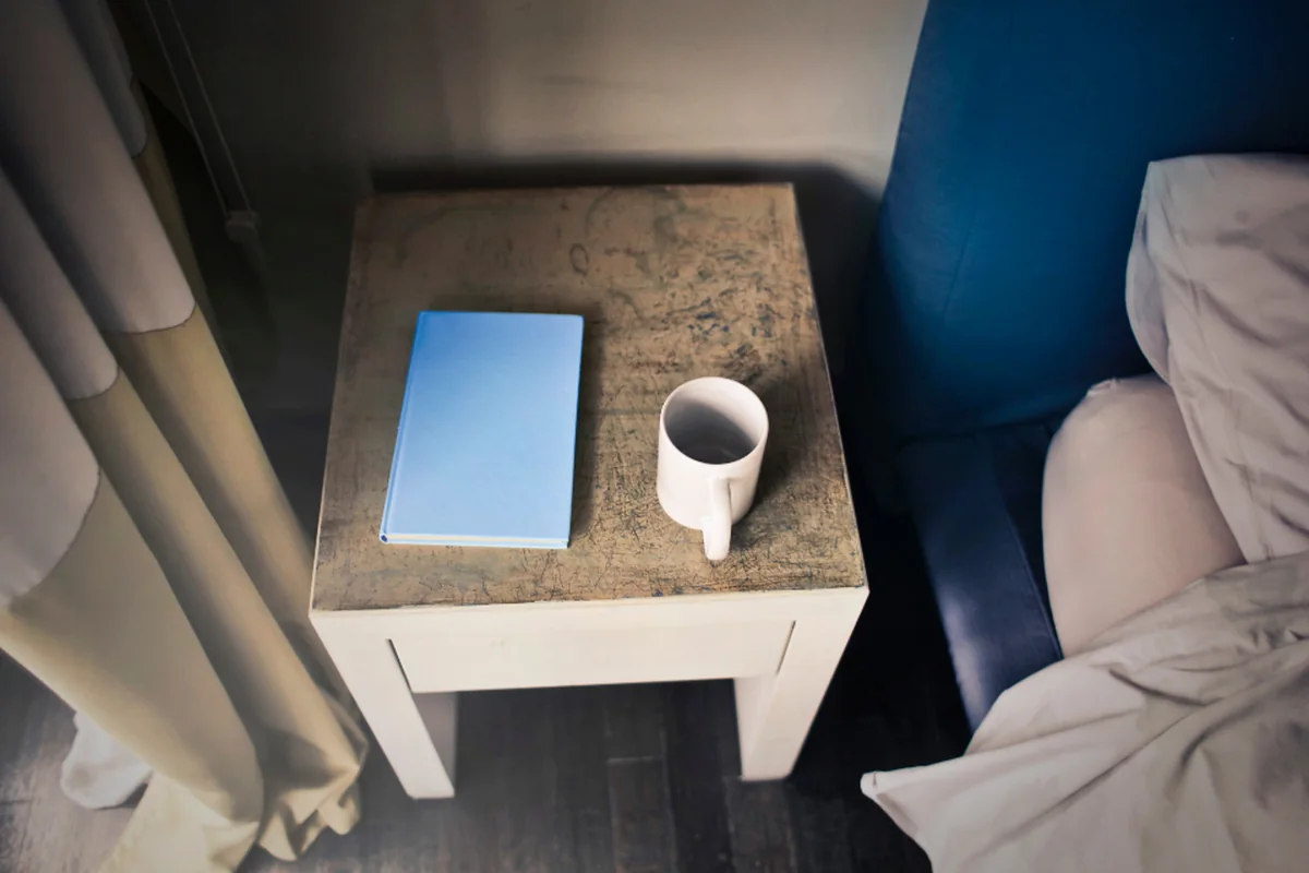 Mesilla de noche de madera con una libreta azul cerrada y una taza blanca, junto a una cama con sábanas claras y una cortina a un lado